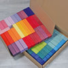 An open cardboard box contains 100 neatly arranged, colorful rectangular blocks. The Large Stepped Pyramid Block Set, organized by color from dark to light shades of blue, green, yellow, orange, red, pink, and purple, provides an excellent tool for developing fine motor skills. The box rests on a light wooden floor.