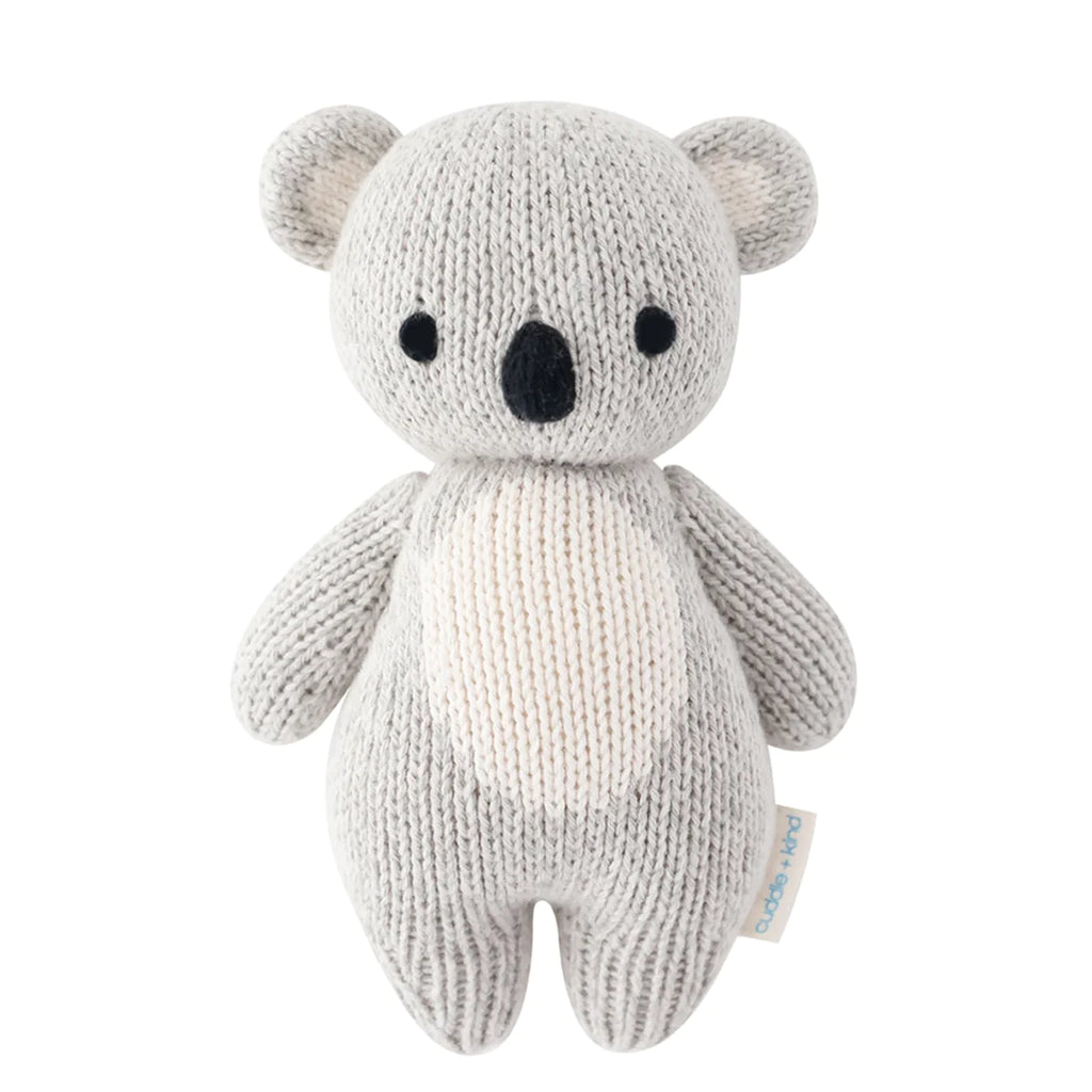 A hand-knit plush Cuddle + Kind Baby Koala toy in neutral colors, standing upright with large black eyes and a small black nose, crafted from Peruvian cotton yarn and isolated on a white background.