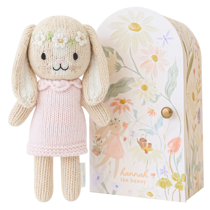 A knitted bunny wearing a pink dress with a floral headband stands next to a decorative box illustrated with flowers and a bunny holding a basket. Handmade in Peru, the box has the text "Cuddle + Kind Tiny Hannah The Bunny" written on it, showcasing fair trade practices.