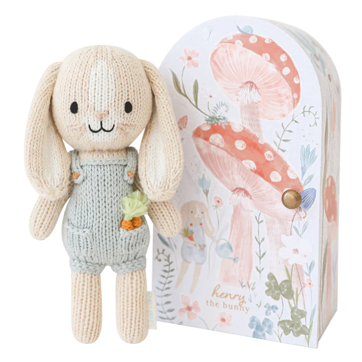 A knitted bunny plush toy, Cuddle + Kind Tiny Henry The Bunny, wearing a light green romper with a tiny carrot in the pocket, stands next to an illustrated cardboard box featuring mushrooms, flowers, and the text "Henry the Bunny." The handmade doll is made from 100% cotton and comes packaged in a whimsically designed box.