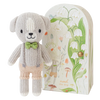 A cute, fair trade Cuddle + Kind Tiny Noah The Dog stands in front of an illustrated book cover. The hand knit cotton doll has a light grey body, a tan face, a green bow tie, and tan pants. The book cover features whimsical botanical illustrations and fairytale elements.