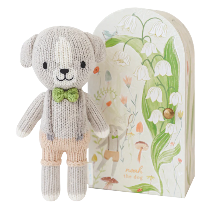 A cute, fair trade Cuddle + Kind Tiny Noah The Dog stands in front of an illustrated book cover. The hand knit cotton doll has a light grey body, a tan face, a green bow tie, and tan pants. The book cover features whimsical botanical illustrations and fairytale elements.