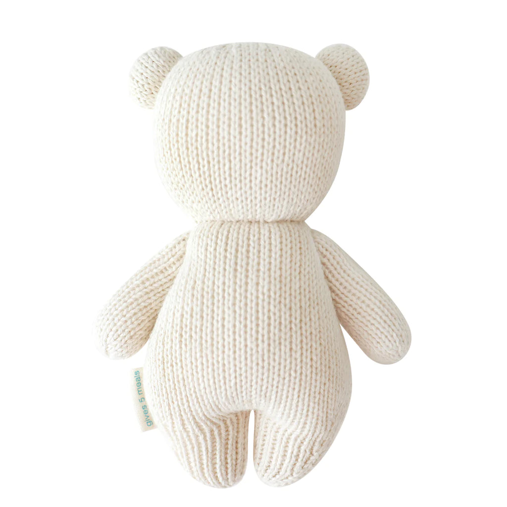 A white, heirloom-quality Cuddle + Kind Baby Polar Bear viewed from the back, hand-knit from natural Peruvian cotton yarn. The bear has a simple design with round ears, a round head, and stubby limbs. A small tag is visible on its left side.