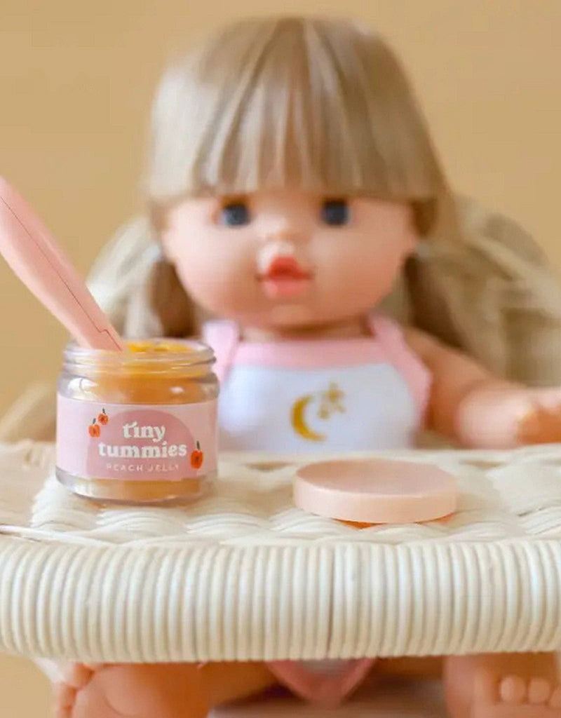 A doll with long blonde hair sits in a woven chair, holding a jar of "Tiny Tummies-Banana Jelly Food" from Tiny Harlow, containing an orange substance. A magic spoon rests in the jar, ready for her next scoop, while the chair features a beige cushion.