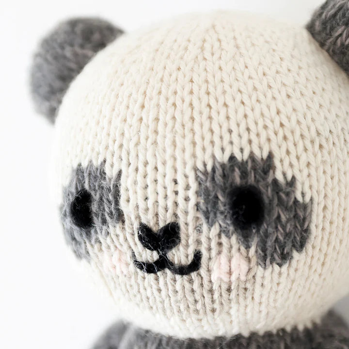 Close-up of a hand-knitted Cuddle + Kind Baby Panda toy with a cream and gray face, featuring black stitched eyes and a smiling mouth. The soft texture of the Peruvian cotton yarn is clearly visible.