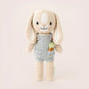 A Cuddle + Kind Tiny Henry The Bunny from the cuddle+kind dolls collection, wearing a light blue romper with a small embroidered carrot on the front. The safety certified bunny has long, floppy ears, a stitched smile, and black eyes against a plain, light-colored background.