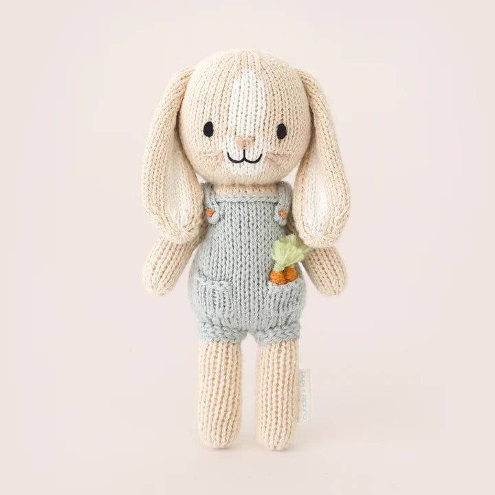 A Cuddle + Kind Tiny Henry The Bunny from the cuddle+kind dolls collection, wearing a light blue romper with a small embroidered carrot on the front. The safety certified bunny has long, floppy ears, a stitched smile, and black eyes against a plain, light-colored background.