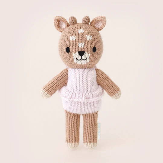 A cute hand-knit toy deer standing upright, featuring a pale brown body, a smiling face, and wearing a cozy white sweater with pink stripes named Cuddle + Kind Tiny Violet The Fawn.