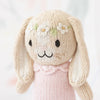 A Cuddle + Kind Tiny Hannah The Bunny with long beige ears, black stitched eyes, and nose. The bunny has small white flower patterns on its head and is wearing a light pink knitted outfit. Made with non-toxic materials, the background is blurred with soft pastel colors.