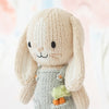 A knitted bunny toy with long ears, named Cuddle + Kind Tiny Henry The Bunny, wears a grey outfit featuring two small knitted carrots in the pocket. Handmade from 100% cotton, the background is softly blurred with pastel colors, giving a gentle and cozy atmosphere.