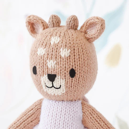 A cute, hand-knit stuffed animal resembling Tiny Violet The Fawn, with a smiling face, prominent ears, and heart-shaped spots on its forehead. It has a soft pink and brown color scheme.