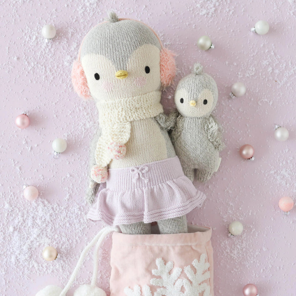 On a pink snowy backdrop, Skylar the Penguin, dressed in a knitted scarf and pink skirt, stands beside a smaller companion. They're nestled in a soft pink bag adorned with snowflakes. Surrounding them are scattered pastel ornaments, each crafted as delightful handmade dolls using sustainable fair trade practices by Cuddle + Kind.