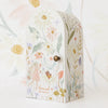 A floral decorative box with an arch-shaped top. The box features illustrations of flowers in pastel colors and a small girl in a pink dress holding hands with a bunny. Embracing fair trade practices, the text on the box reads “Cuddle + Kind Tiny Hannah The Bunny” in lowercase letters.