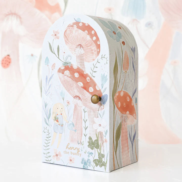 A whimsical illustration on a pastel-colored, arched-top cardboard box features a bunny holding a flower amidst tall, red-capped mushrooms with white spots and various delicate plants. The text "Cuddle + Kind Tiny Henry The Bunny" is inscribed on the box. Inspired by cuddle+kind dolls, this design evokes hand-knit charm.