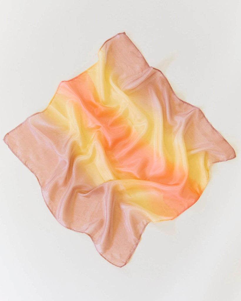 A charming, soft square silk scarf named Sarah's Silk Enchanted Playsilk - Desert features a pastel gradient of pink, yellow, and orange hues as it lies casually on a plain white background. Resembling playsilks, the fabric invites imaginative play with its smooth yet slightly crinkled appearance.