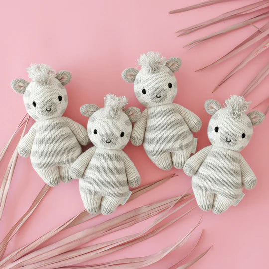 Four adorable hand-knit Cuddle + Kind Baby Zebra dolls in a row on a pink background, surrounded by soft pink palm fronds. Each giraffe has a sweet smile and tufted hair.