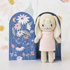 A Cuddle + Kind Tiny Hannah The Bunny doll wearing a light pink dress stands in front of an open book-shaped background. The backdrop features dark blue panels adorned with colorful flowers, a crescent moon, and stars. The setting is light, with more floral designs in the background—showcasing our commitment to fair trade practices.