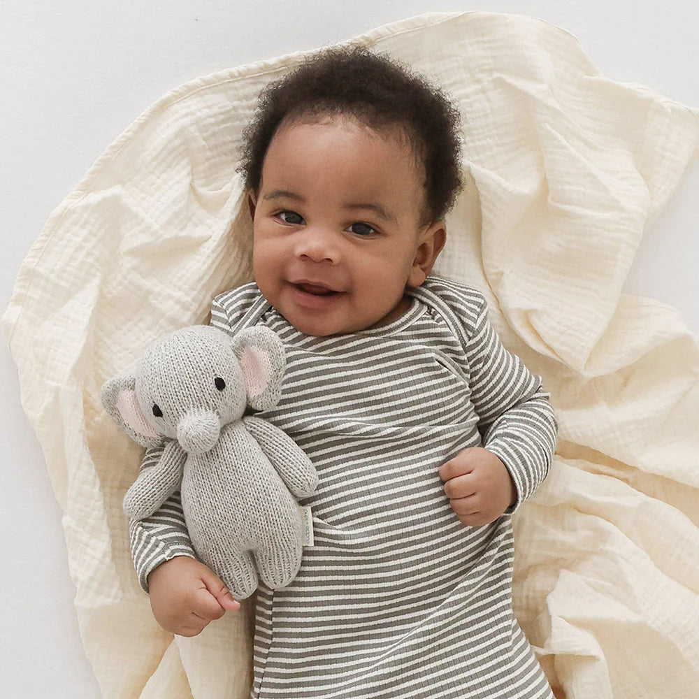 A baby wearing a gray and white striped onesie lies on a cream-colored blanket, smiling at the camera while holding a Cuddle + Kind Baby Elephant made from soft Peruvian cotton yarn.