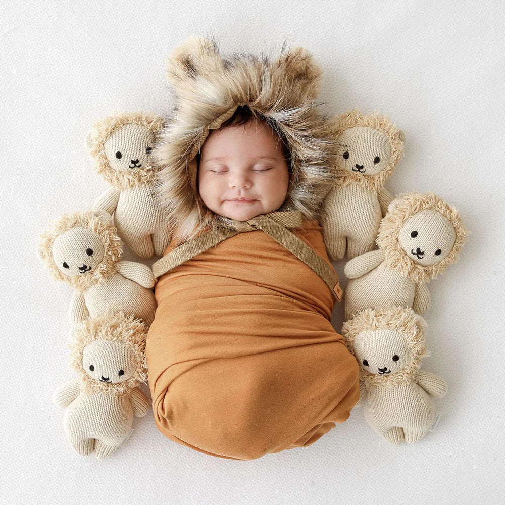 A newborn baby wrapped in an orange swaddle sleeps peacefully, surrounded by six small teddy bears against a white backdrop. The baby wears a hand-knit furry hat with ear flaps made of Cuddle + Kind Baby Lion.