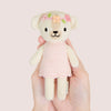 A hand holding an adorable plush toy dog named Tiny Charlotte from Cuddle + Kind, dressed in a pink, hand-knit dress. The dog features embroidered flowers on its head and a smiling face, crafted from non-toxic yarn, set against a soft beige background.