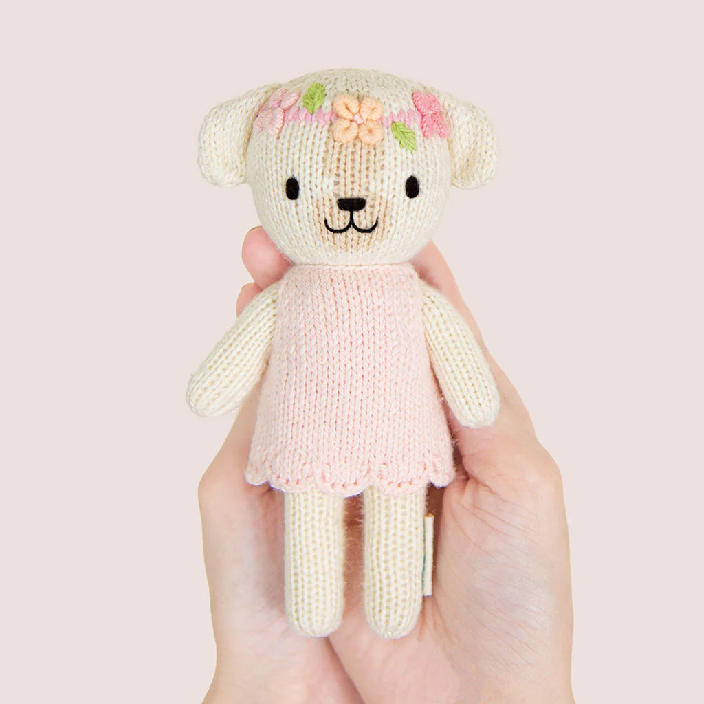 A hand holding an adorable plush toy dog named Tiny Charlotte from Cuddle + Kind, dressed in a pink, hand-knit dress. The dog features embroidered flowers on its head and a smiling face, crafted from non-toxic yarn, set against a soft beige background.