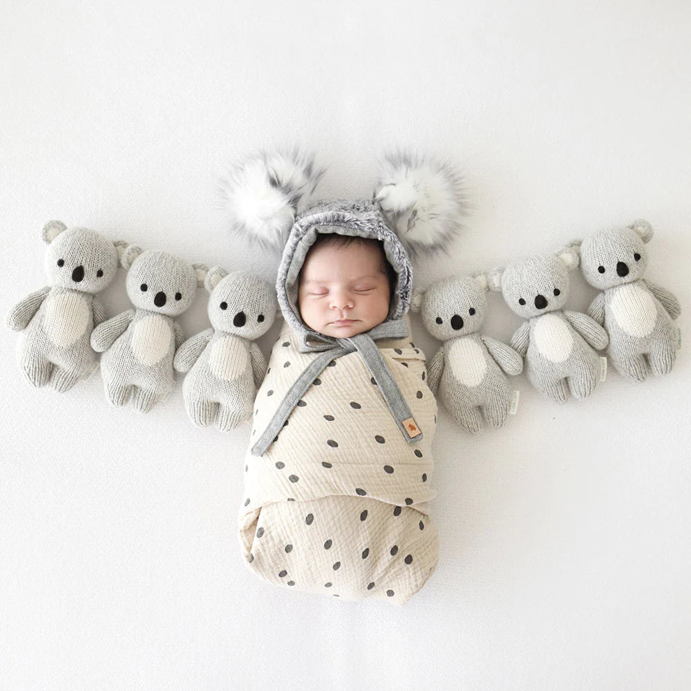 A sleeping newborn baby swaddled in a light polka-dot wrap, wearing a cozy mouse-eared cap, flanked by rows of hand-knit baby animals, crafted from Cuddle + Kind Baby Koala yarn