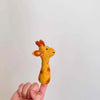 A Giraffe Finger Puppet, featuring a charming brown and yellow needle-felted design, is perched on an adult's index finger against a plain white background. This handcrafted wool puppet boasts small ears, two horn-like structures atop its head, and a cheerful smiling expression.