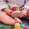 A baby wearing a white outfit with a floral pattern sits on a textured surface, holding a Mini Rolling Wheel with colorful bars and wooden beads. Several colorful balls are positioned near the baby's feet. The focus is on the baby's hands and the toy.