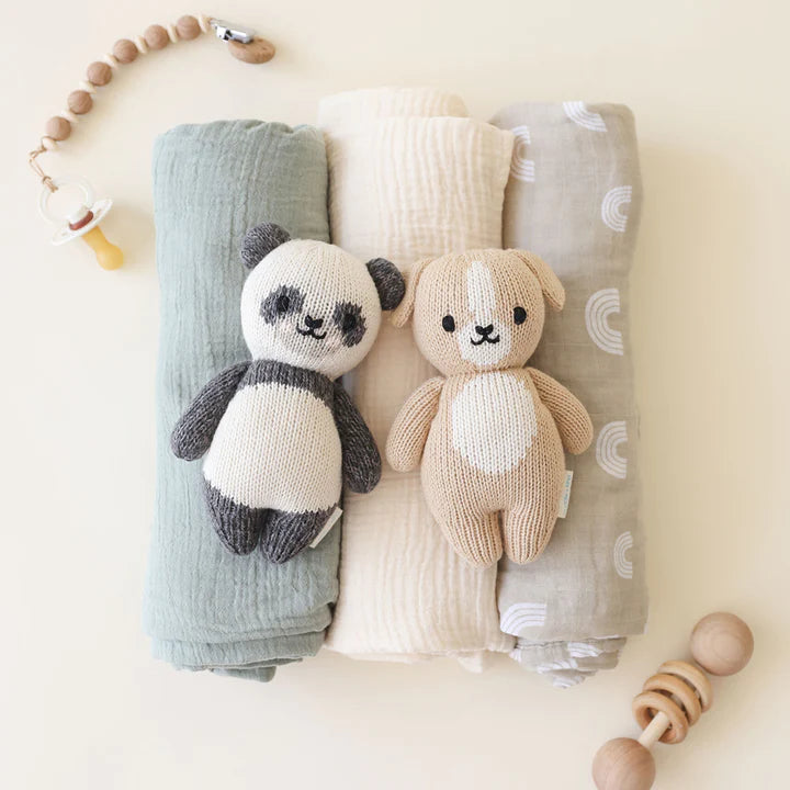 Two hand-knit Cuddle + Kind Baby Panda dolls, made from Peruvian cotton yarn, lying on folded baby blankets with a light wooden beads toy and pacifier next to them