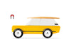 The Candylab Cotswold is a toy vehicle featuring a bright yellow body, a white roof, and a wooden surfboard on top. It has black wheels with "CANDYCAR" written on them. Near the rear of the car, there is a red and black magnet symbol indicating its magnetic feature for comfortable travel. The car is set against a white background.