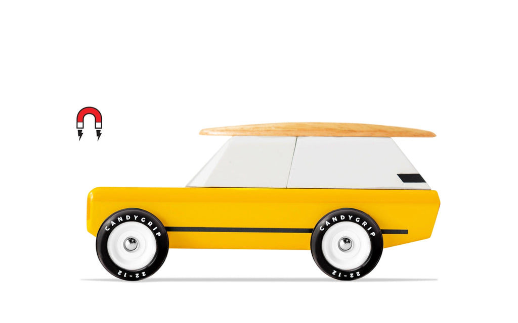 The Candylab Cotswold is a toy vehicle featuring a bright yellow body, a white roof, and a wooden surfboard on top. It has black wheels with "CANDYCAR" written on them. Near the rear of the car, there is a red and black magnet symbol indicating its magnetic feature for comfortable travel. The car is set against a white background.