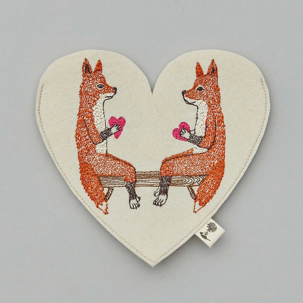 Embroidered Coral & Tusk Smitten Foxes Felt Envelope depicting two orange foxes sitting on a bench, each holding a pink heart, set against a light grey background.