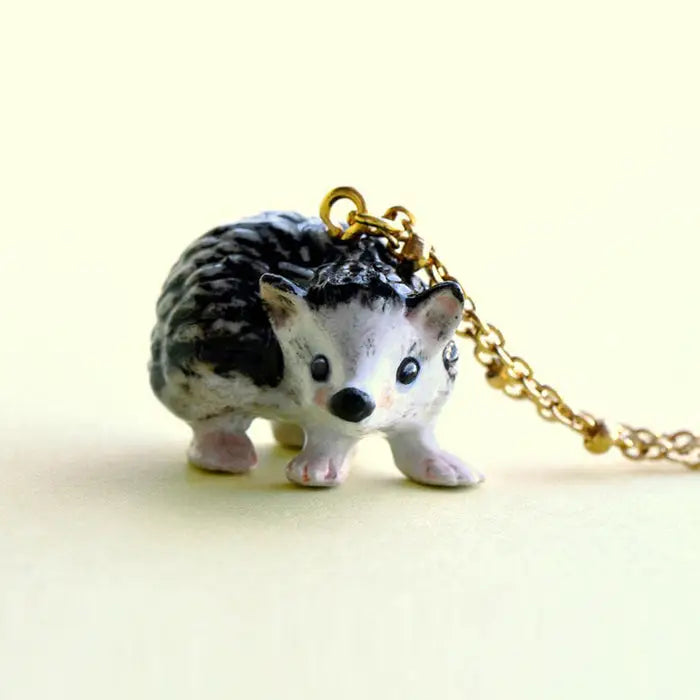 A close-up photo of a Hedgehog Necklace on a 24k gold-plated chain. The hand-painted pendant is black and white with detailed textures to mimic the appearance of a hedgehog, set against a light yellow background.