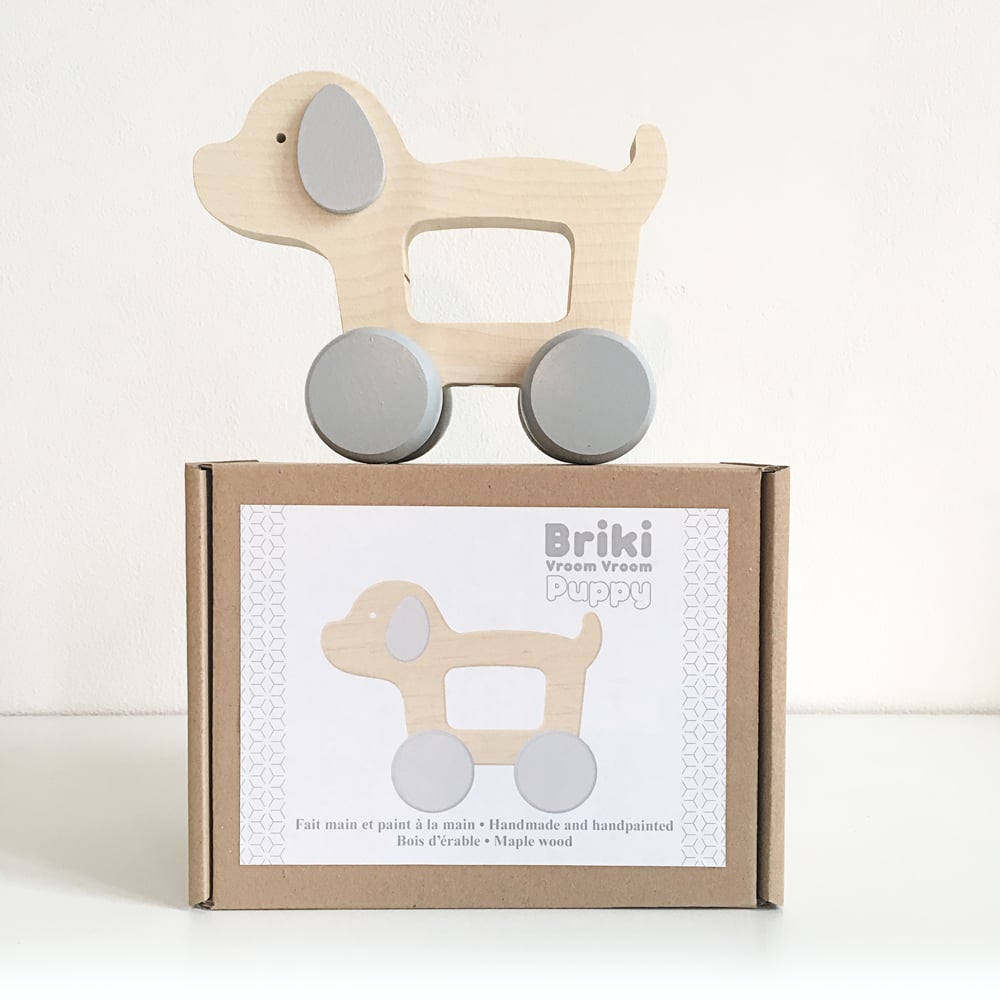 A Wooden Puppy Push Toy with gray wheels stands on top of its packaging box. The box features an illustration of the toy and text in both French and English, indicating that it is handmade, hand-painted, and made from maple wood. This delightful toy enhances fine motor skills for young children.