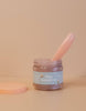 An open jar with a blue label displaying "Tiny Tummies - Coconut Yogurt" from Tiny Harlow is accompanied by a pink spoon protruding from it. The jar sits next to its matching pink lid against a beige backdrop.