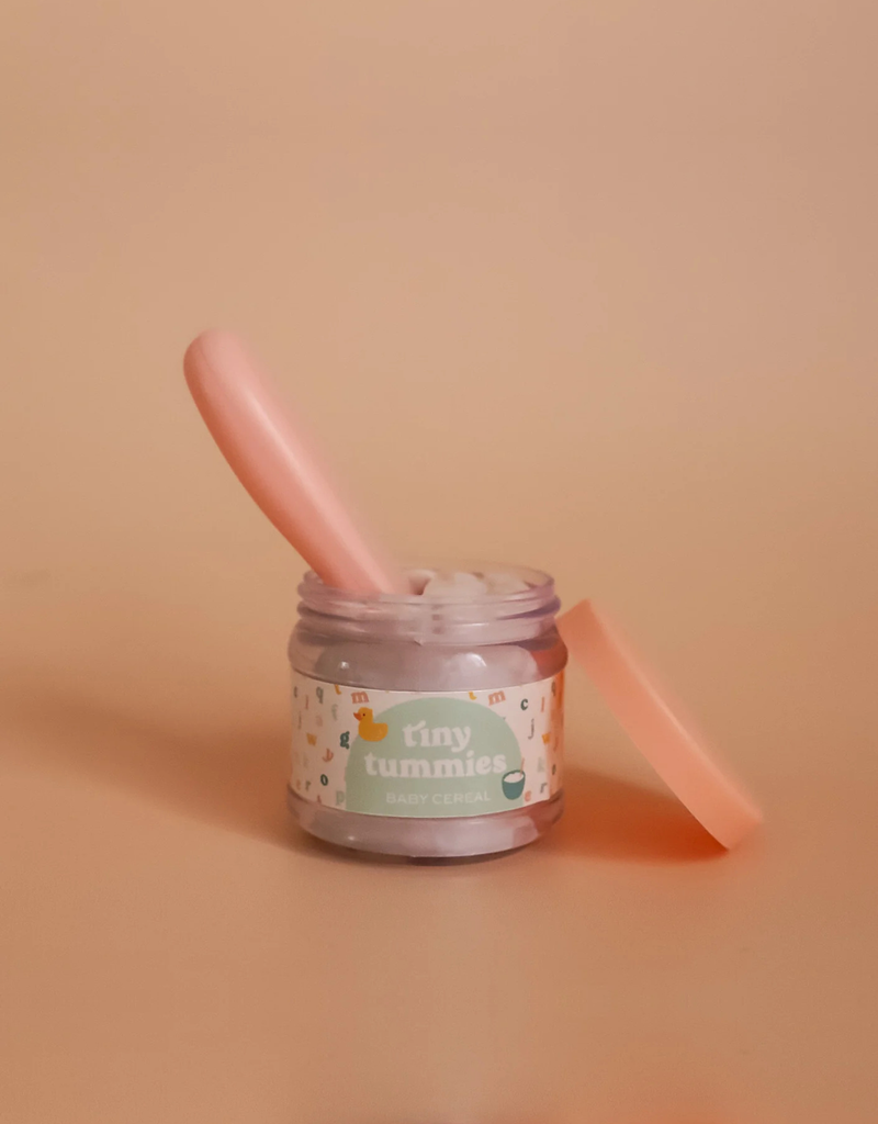 A small jar labeled "My First Tiny Tummies gift Set - Milk and Cereal" is open, with a light pink spoon resting inside. The enchanting jar displays whimsical designs adorned with tiny illustrations, and the lid sits off to the side against a soft beige background.