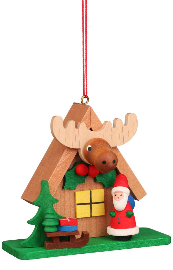 The Christian Ulbricht Ornament - Santa with Elk House features a charming wooden house adorned with an elk head above the window. Next to it, there's a Santa figurine standing beside a Christmas tree and a sleigh filled with presents, all expertly crafted in Germany. The ornament hangs elegantly from a bright red string.