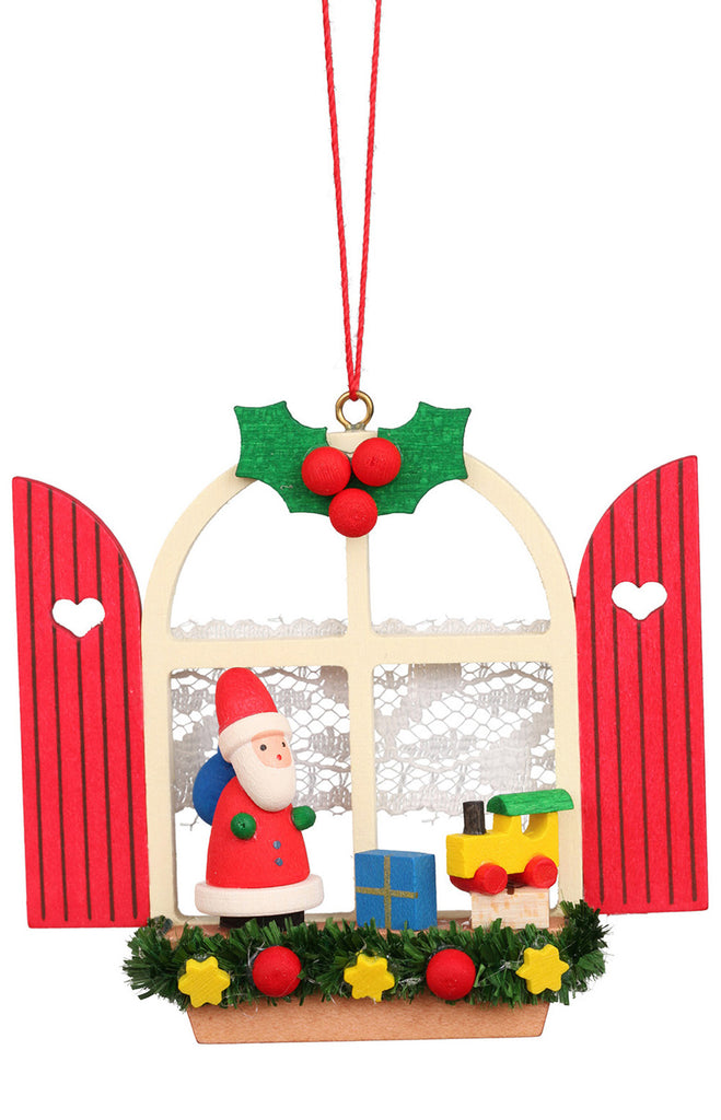 The Christian Ulbricht Ornament - Window With Santa is a festive Christmas decoration crafted in Germany, showcasing a wooden Santa figure next to a small train and gift. This charming hanging ornament is beautifully framed by a window with lace curtains, red shutters, holly leaves, and berries, and features greenery and yellow stars below.