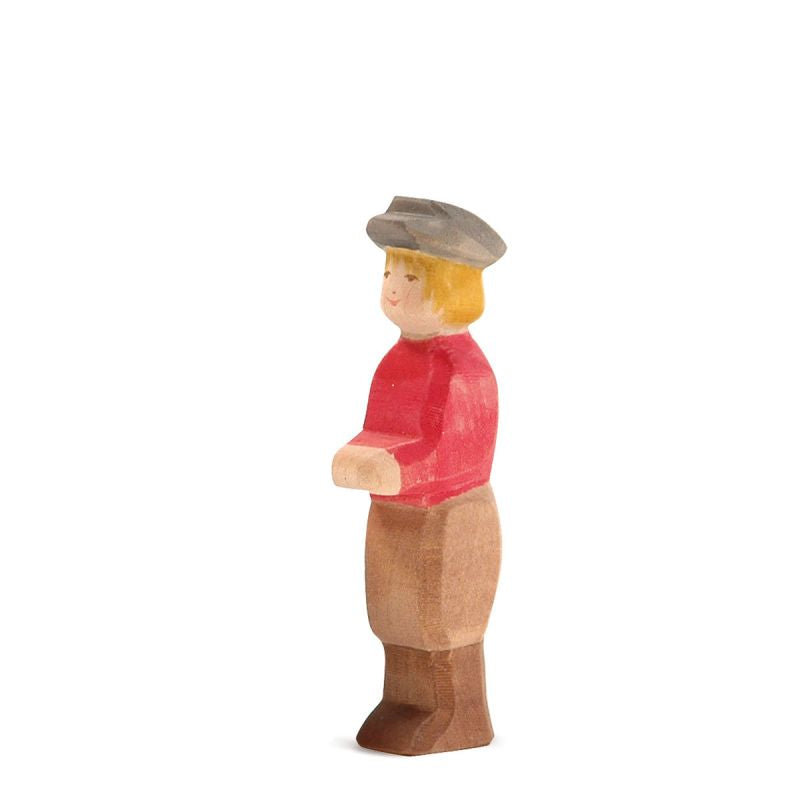 The Ostheimer Son, Gray Hat is a wooden figurine featuring a person with short light brown hair, donning a red shirt, brown pants, and a gray hat. The figure stands upright and appears hand-painted with simple facial features and exudes a rustic, handcrafted charm typical of Ostheimer toys.