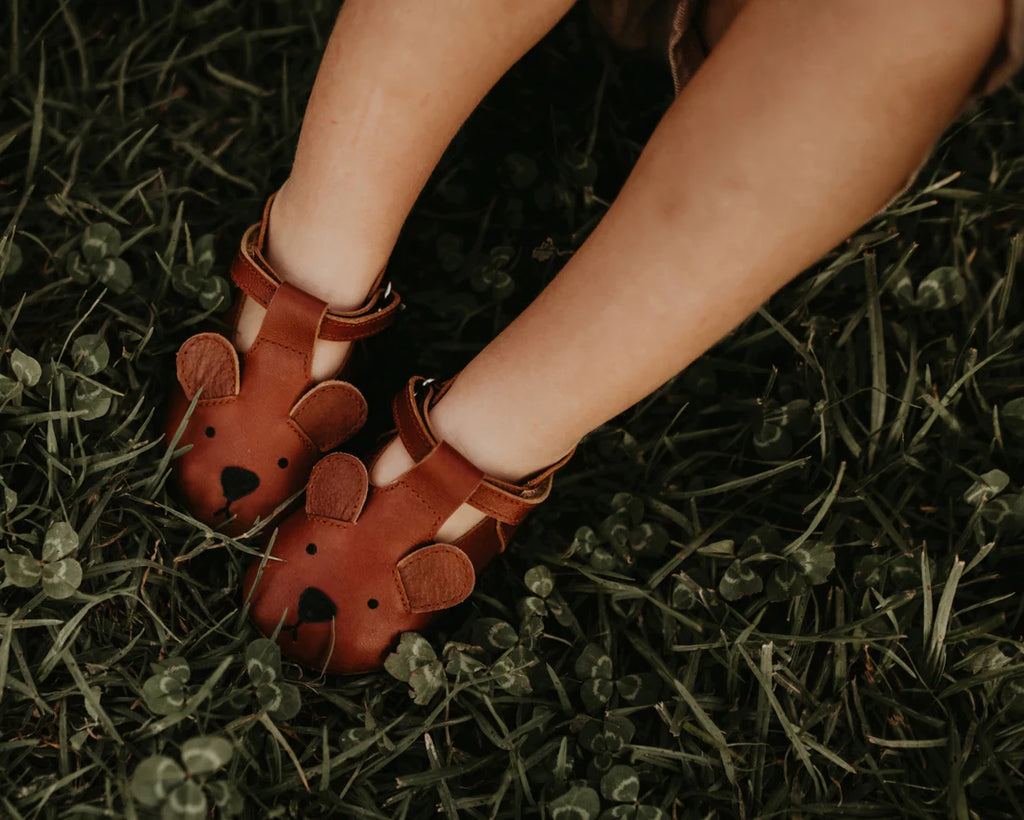 A child's feet in Donsje Baby Bear Shoes, resting on green grass filled with clovers.