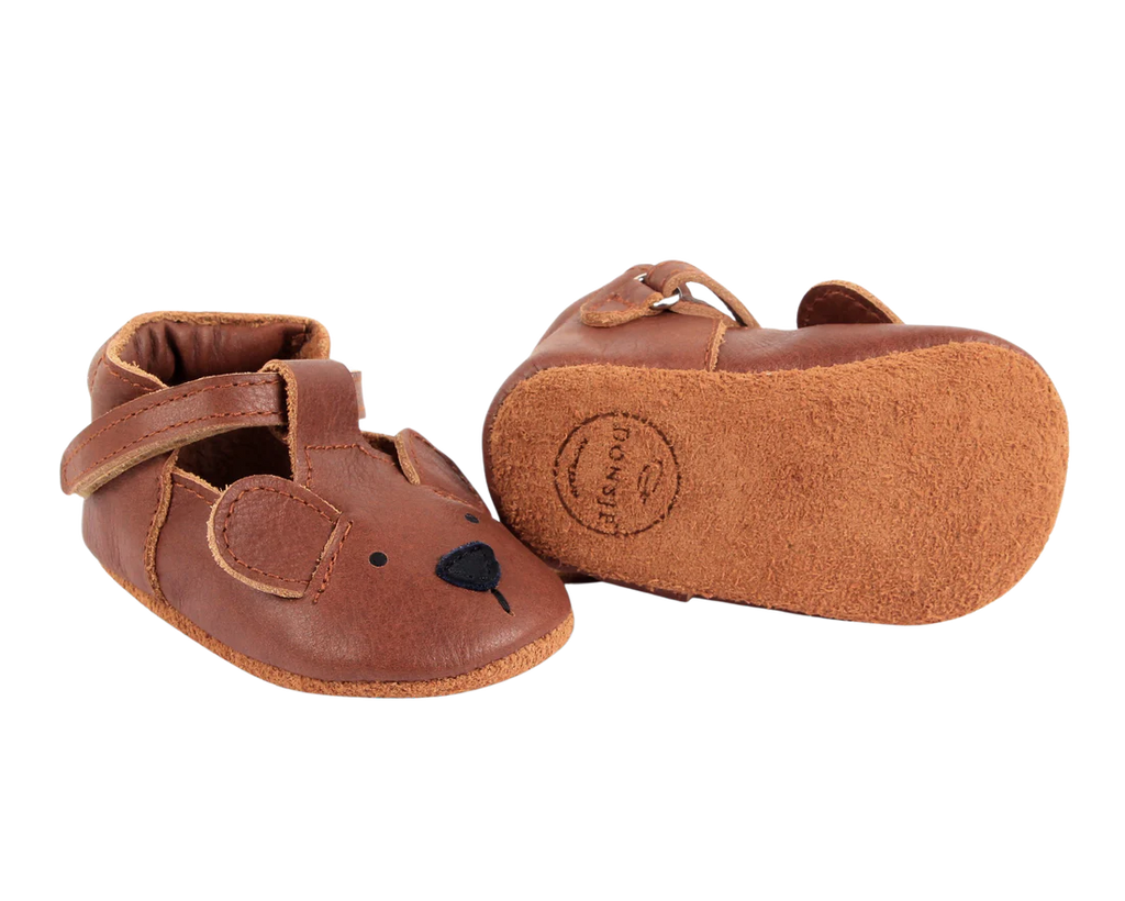 A pair of Donsje Baby Bear Shoes with a bear face design and a Velcro fastening strap, displayed against a transparent background.