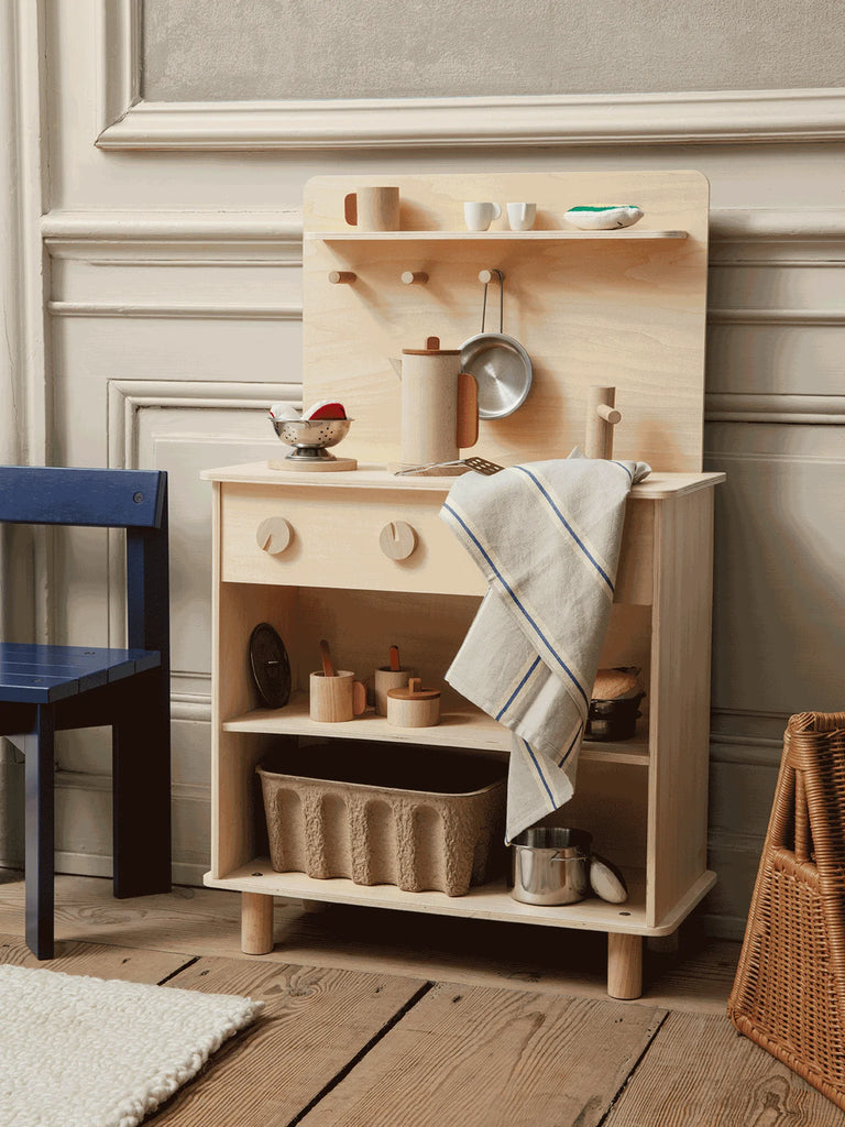 A Ferm Living Toro Play Kitchen set against a wall next to a blue chair, accessorized with miniature utensils, pots, and a striped towel hanging on the side.