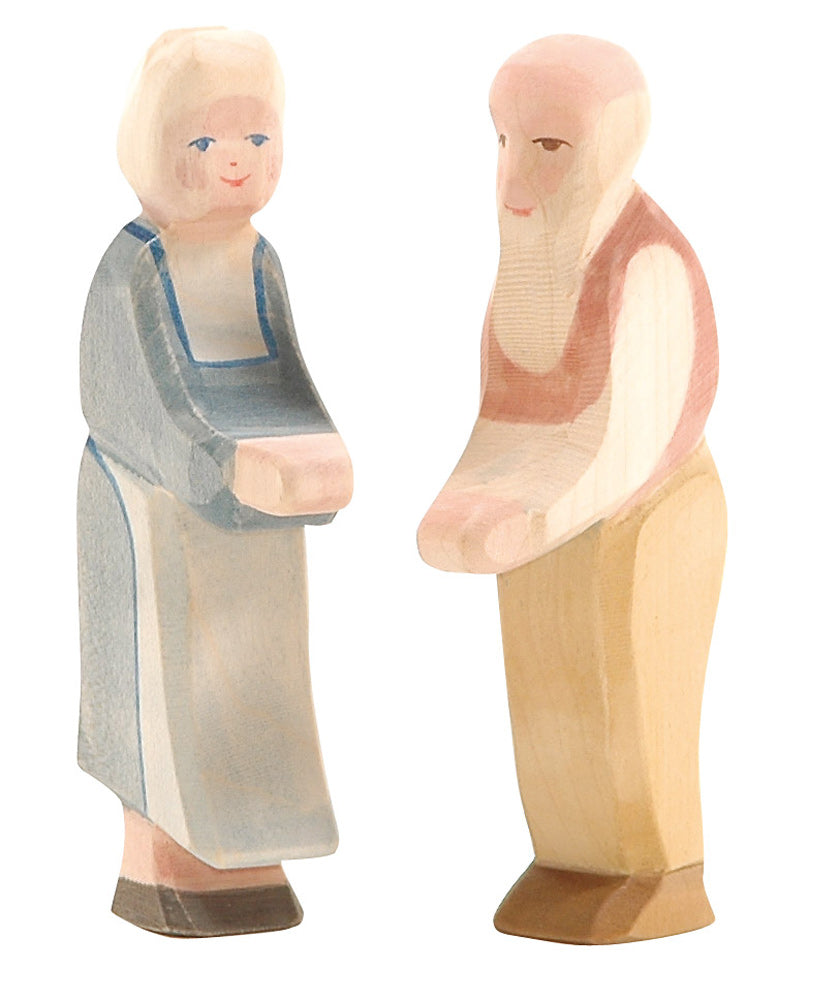 The Ostheimer Grandmother figurine depicts an elderly woman with gray hair wearing a blue dress. Accompanying her is an elderly man figure, notable for his white beard and bald head, dressed in a vest and pants. Both handcrafted wooden toys are hand-painted, facing forward, and posed as if holding objects.