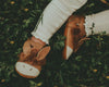 Close-up of a child's feet in Donsje Baby Donkey Shoes with a velcro fastening strap, on a bed of small green plants and flowers.