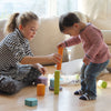 A young girl and a toddler play with the colorful, non-toxic Grimm's Tree Slices Building Blocks on a hardwood floor. The girl, wearing a striped shirt and polka dot leggings, helps the toddler, who is dressed in a striped long-sleeve shirt and jeans, stack the building blocks. Bright, natural light fills the room.