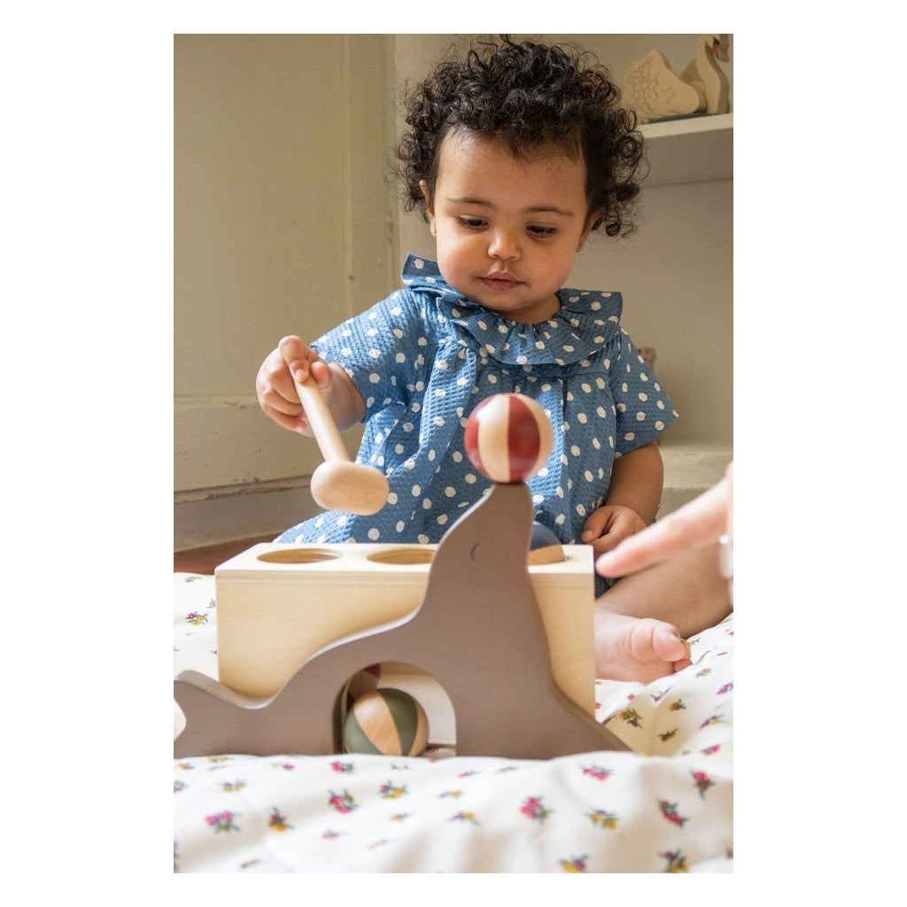 A toddler with curly hair, wearing a blue polka dot dress, plays with a Wooden Hammer Game - Sea Lion made from FSC-certified wood, while seated on a floral mat.