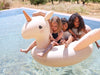 Three children happily sitting on an Inflatable Unicorn Float with handles on the neck in a pool on a sunny day, surrounded by trees.