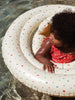 A toddler with curly hair wearing a red swimsuit with white polka dots sits in an Inflatable Baby Swim Ring - Multi Hearts. The durable PVC ring, which complies with EN 13138-3:2021 standards, gently floats on the shallow water's surface as the toddler looks to the side while holding onto it.