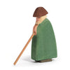 A handcrafted wooden figurine of a person wearing a green cloak and a brown hat, leaning on a wooden walking stick. The figure, reminiscent of Ostheimer Shepherd With Staff, has a simple, carved design with minimal facial features and stands against a plain white background.