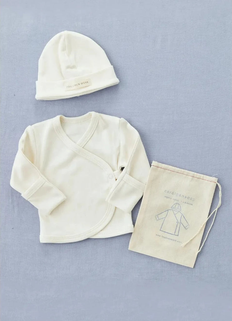 A neatly arranged display of a cream-colored Baby Cap & Cardigan made from organic cotton. The set includes a long-sleeved wrap-around cardigan with side snap closures, a matching baby hat, and a small beige drawstring bag with an illustration and text. The items are on a light blue background.